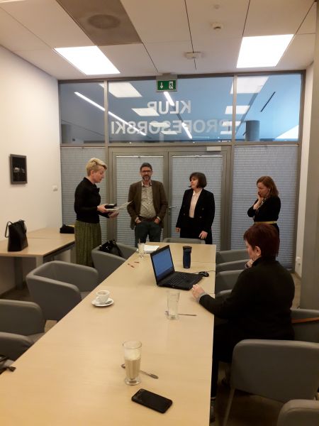 4 osoby stoją i rozmawiają w klubie profesorskim w bibliotece WFPiK, jedna osoba siedzi przy stole, przed nią stoi otwarty laptop