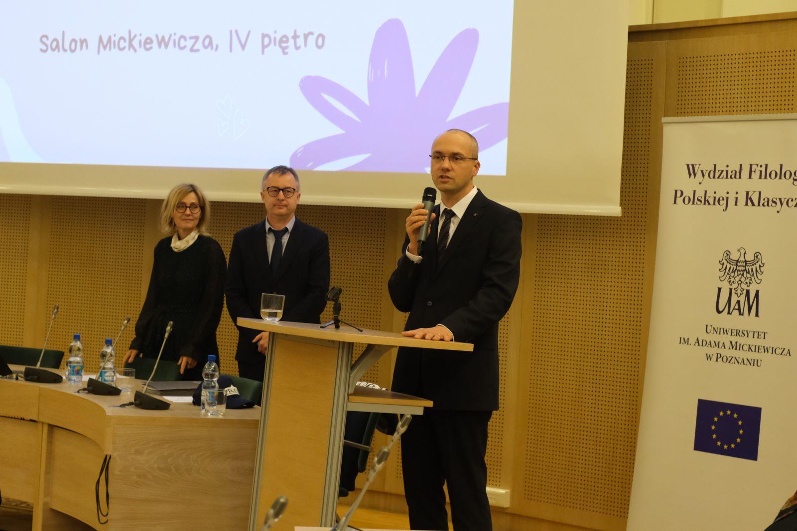 Prof. Krzysztof Skibski stoi przy mównicy i przemawia do mikrofonu, za nim stoją prof. Tomasz Mizerkiewicz i prof. Krystyna Pieniążek-Marković.