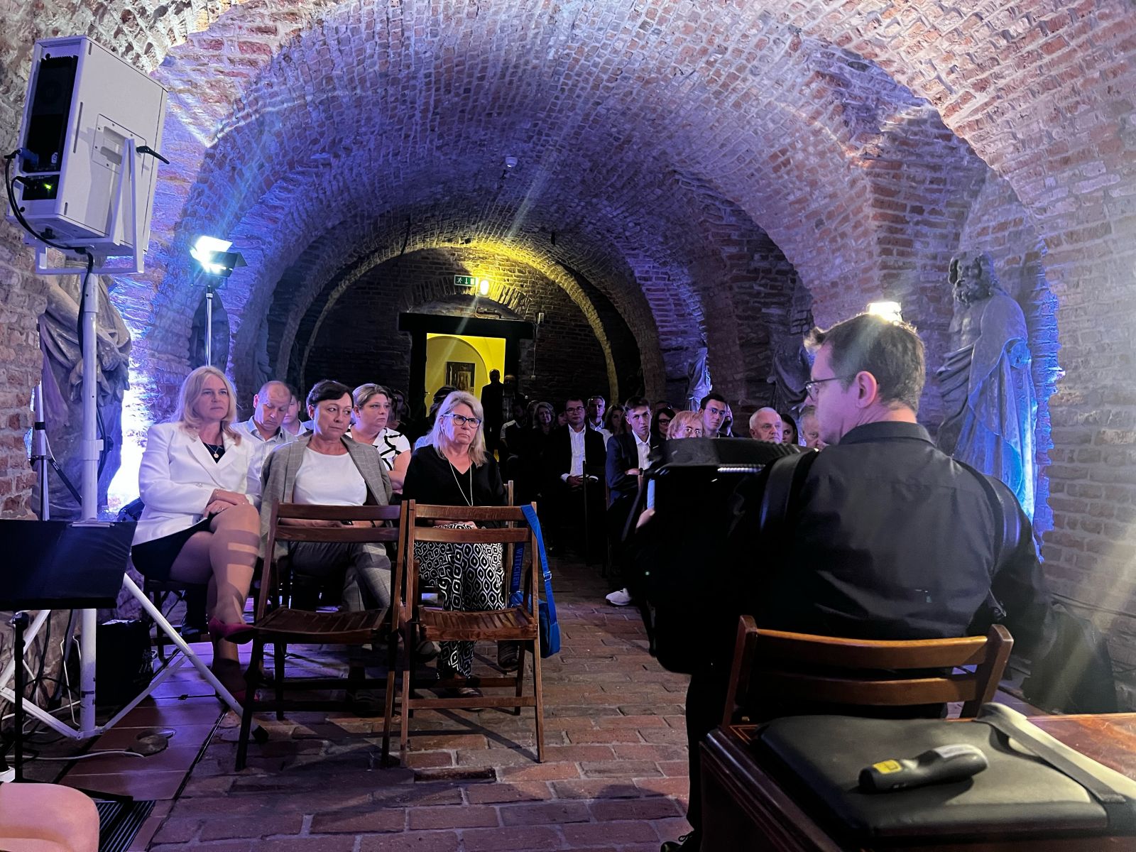 Widok na pomieszczenie z cegieł od strony podwyższenia, na którym siedzi mężczyzna z akordeonem, przed nim publiczność na drewnianych krzesłach.