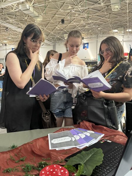 Trzy młode kobiety trzymają w dłoni broszurę z fioletową okładką, drugą rękę przytykają do brody w wyrazie skupienia.