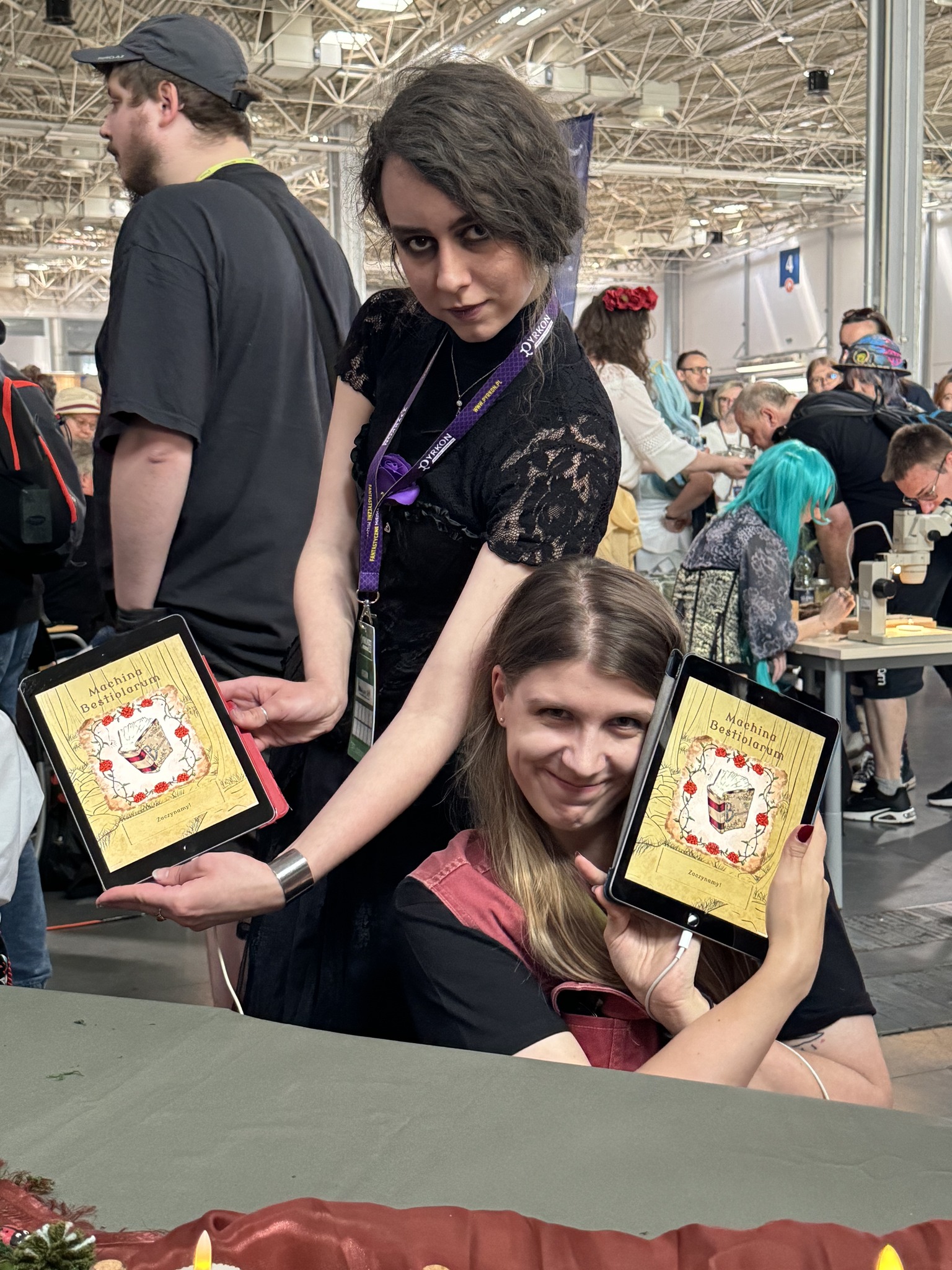 Studentki prezentują tablety z aplikacją do tworzenia fantastycznych chowańców, za nimi widać przechodzących uczestników festiwalu Pyrkon.