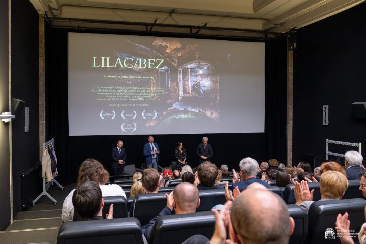 Twórcy filmu stoją pod ekranem kinowym, na którym wyświetlony jest plakat filmowy, przed nimi widać zgromadzoną publiczność siedzącą w sali kinowej.