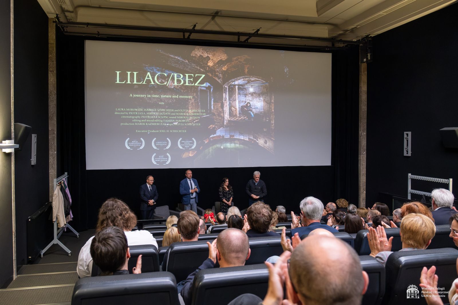Twórcy filmu stoją pod ekranem kinowym, na którym wyświetlony jest plakat filmowy, przed nimi widać zgromadzoną publiczność siedzącą w sali kinowej.