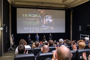 Twórcy filmu stoją pod ekranem kinowym, na którym wyświetlony jest plakat filmowy, przed nimi widać zgromadzoną publiczność siedzącą w sali kinowej.