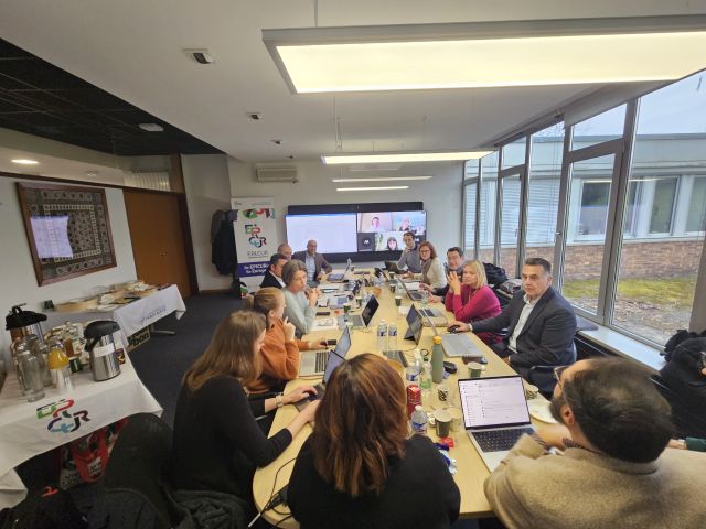 Uczestnicy spotkania Managemenet Board siedzą przy stole roboczym, większość ma przed sobą otwarte laptopy, w głębi pomieszczenia na rzutniku widać konferencję w Microsoft Teams.