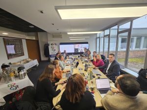 Uczestnicy spotkania Managemenet Board siedzą przy stole roboczym, większość ma przed sobą otwarte laptopy, w głębi pomieszczenia na rzutniku widać konferencję w Microsoft Teams.