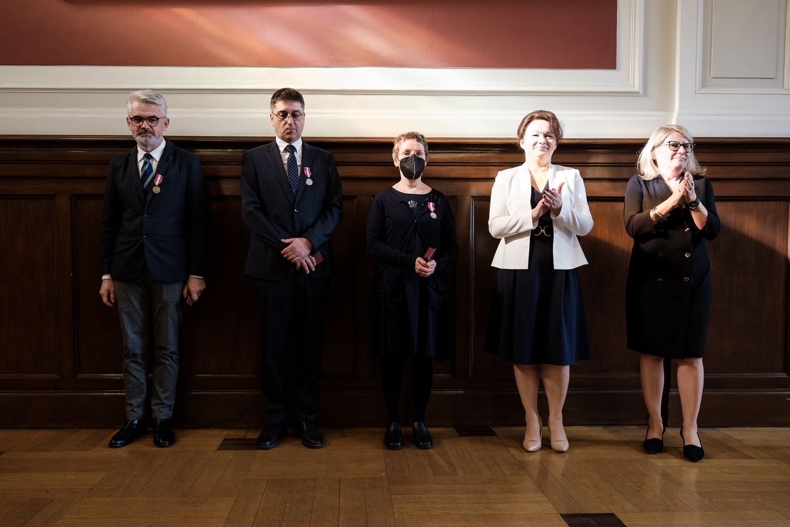 Na tle ściany wyłożonej drewnem stoi pięć osób, trzy z nich na piersi mają medal, dwie klaszczą.