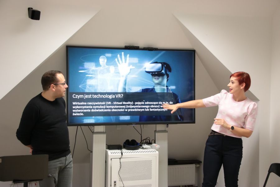 Na dużym ekranie telewizora wyświetlana jest prezentacja o VR, przed ekranem dwie osoby objaśniają szczegóły.