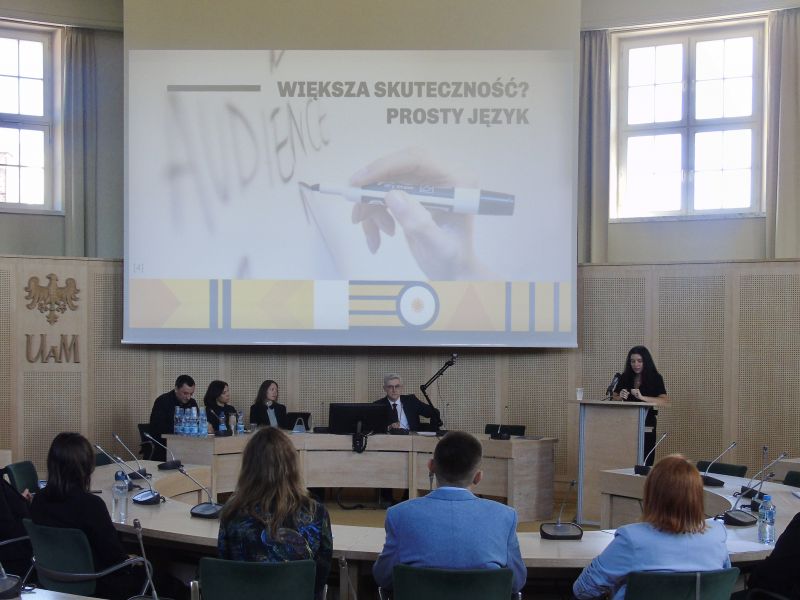 W Salonie Mickewicza za stołem prezydialnym siedzą 4 osoby, jedna osoba stoi przy mównicy i przemawia, na ekranie wyświetla się prezentacja, widać również plecy osób na widowni.