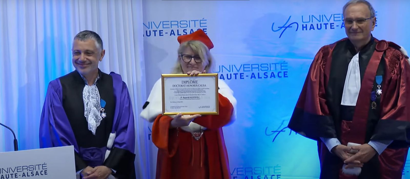 Ubrana w szaty rektorskie Rektor UAM Bogumiła Kaniewska trzyma w dłoniach dyplom doktora honoris causa, prezentując go publiczności, z jej lewej i prawej strony stoją dwaj mężczyźni w togach.