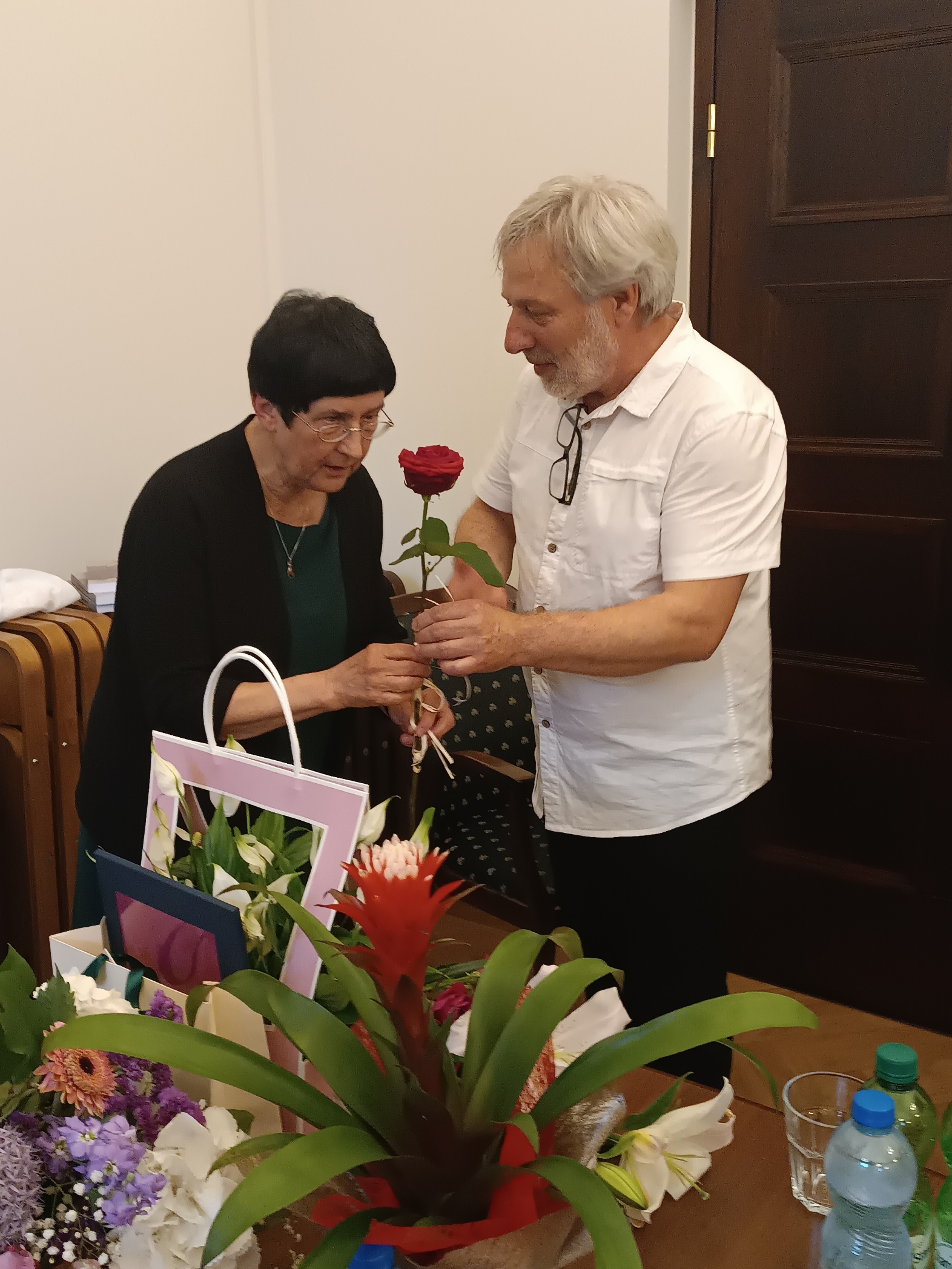 Prof. Zbigniew Kopeć wręcza prof. Sienkiewicz czerwoną różę, mówiąc coś do jubilatki. Przed nimi na stole leży mnóstwo kwiatów i prezentów.