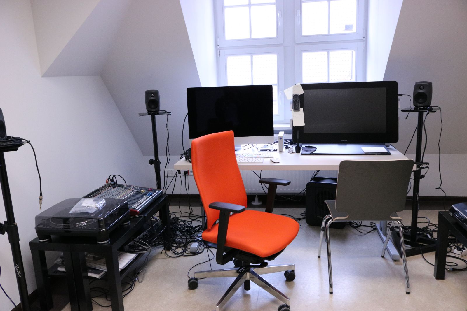 Zdjęcie biurka z dwoma dużymi ekranami komputera, przed którym stoi pomarańczowy fotel biurowy, a obok różny sprzęt do nagrywania i odtwarzania dźwięku, sporo zwojów kabli, mikrofony itp.