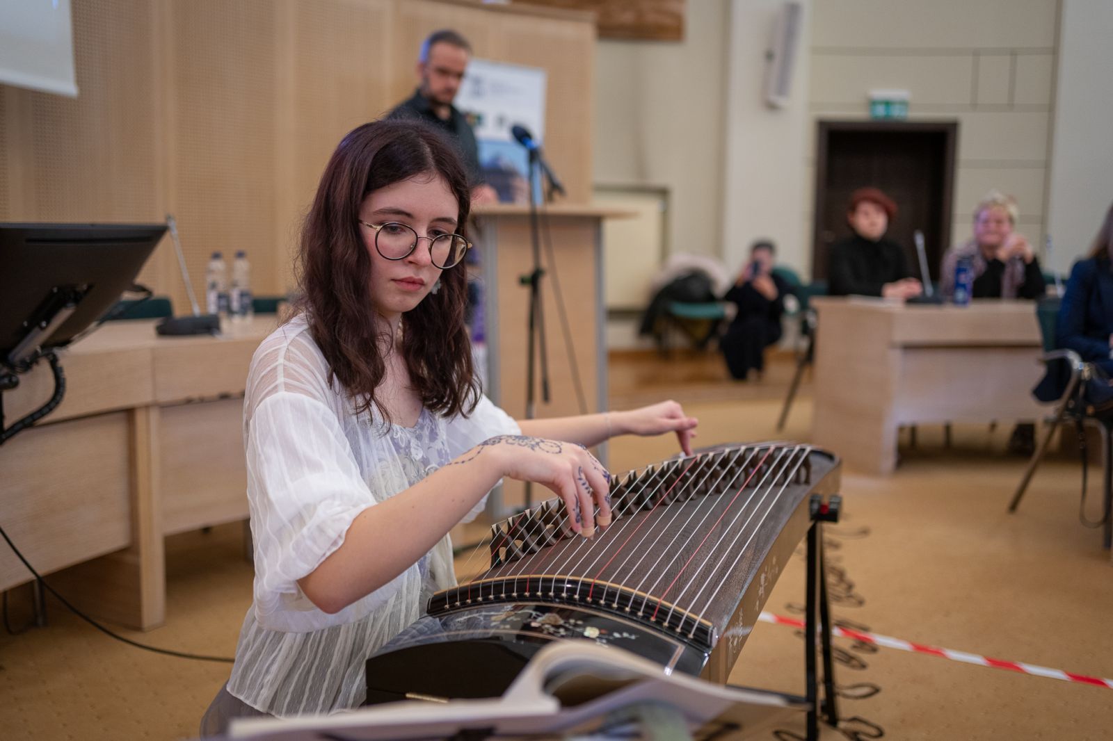 Młoda kobieta siedzi przy tradycyjnym chińskim instrumencie, przypominający długi stół ze strunami i gra na nim.