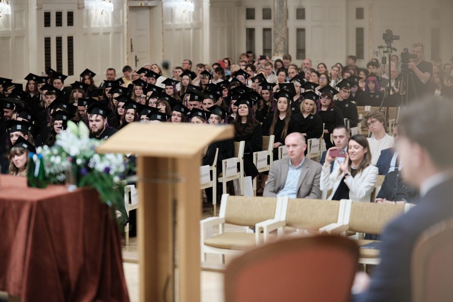 Uczestnicy absolutorium ubrani w togi i czapki absolwenckie siedzą w rzędach w Auli Uniwersyteckiej. 