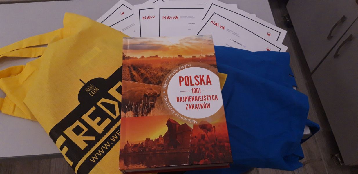 Na stoliku leżą certyfikaty uczestnictwa w kursie języka polskiego, torby z logotypem Fredry 10 i książka 
