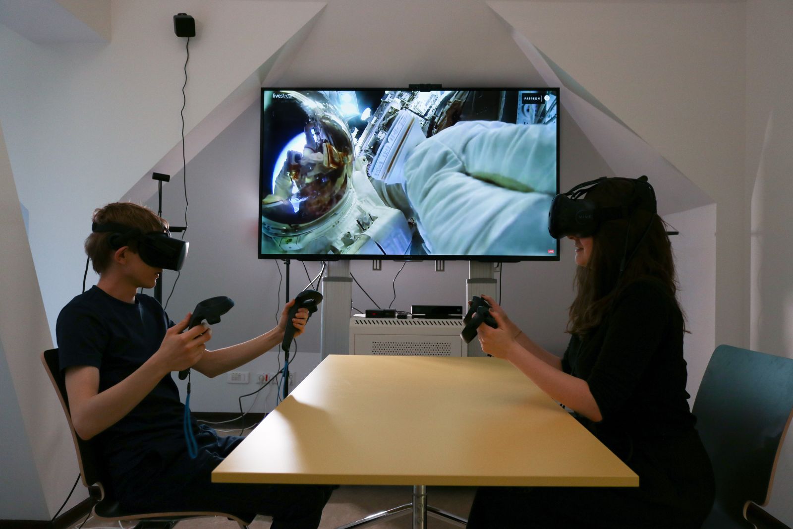 Dwie osoby w okularach VR siedzą przy stole, w rękach trzymają specjalne kontrolery, za nimi na ekranie wyświetla się kadr z kosmonautą szybującym w kosmosie.