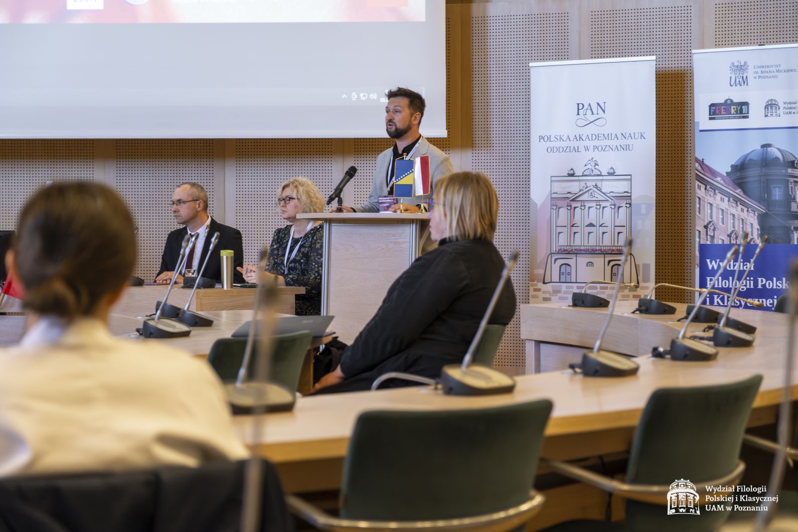 Dr hab. Patryk Borowiak przemawia przy mównicy. Obok niego widać część stołu prezydialnego oraz część zgromadzonej publiczności.