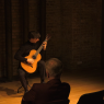 Wykonawca - Andrzej Grygier - siedzi na krześle w Sali Teatralnej, w przytłumionym świetle, grając na gitarze. Na pierwszym planie widać ciemne sylwetki publiczności z pierwszego rzędu.