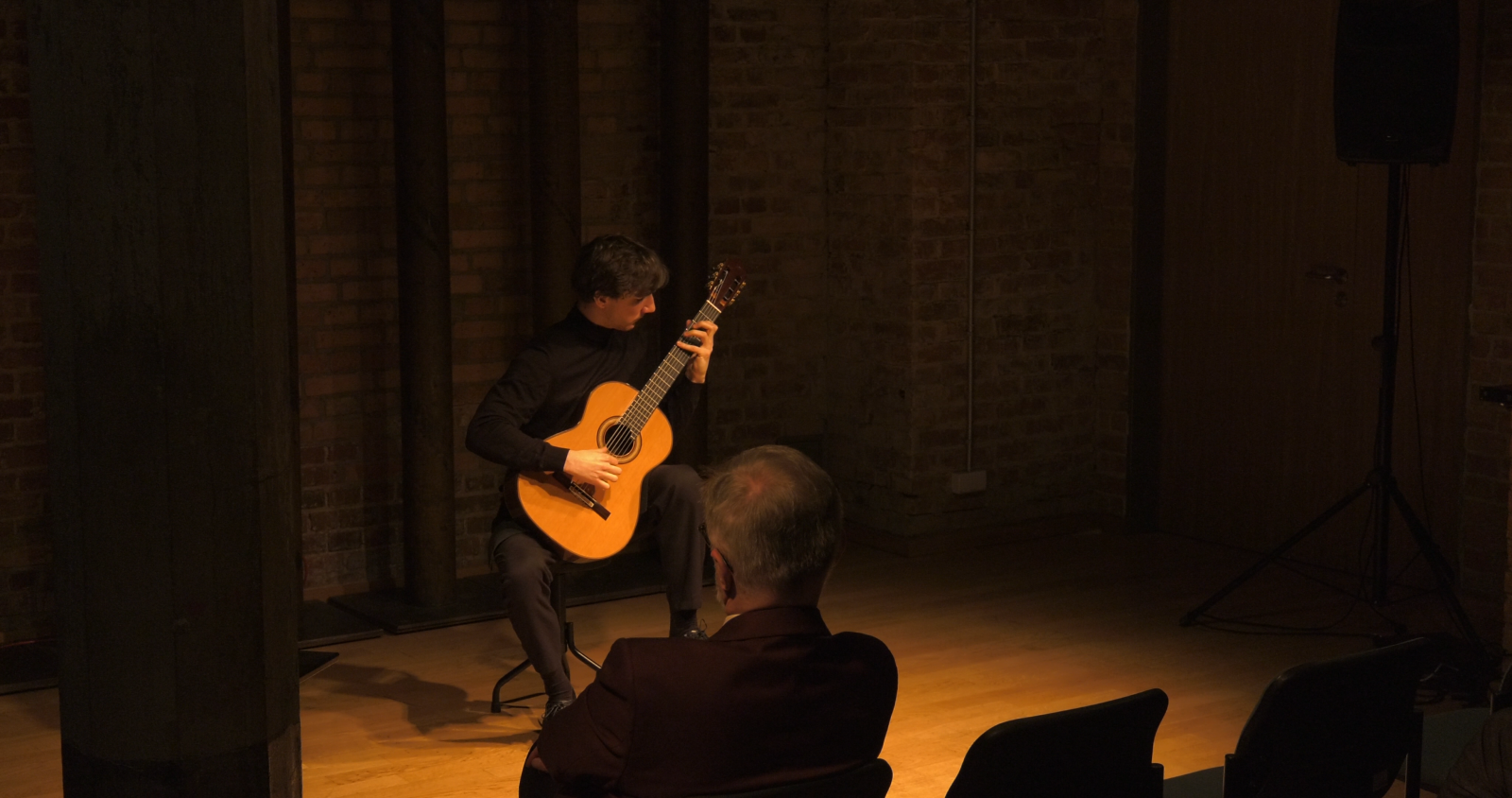 Wykonawca - Andrzej Grygier - siedzi na krześle w Sali Teatralnej, w przytłumionym świetle, grając na gitarze. Na pierwszym planie widać ciemne sylwetki publiczności z pierwszego rzędu.