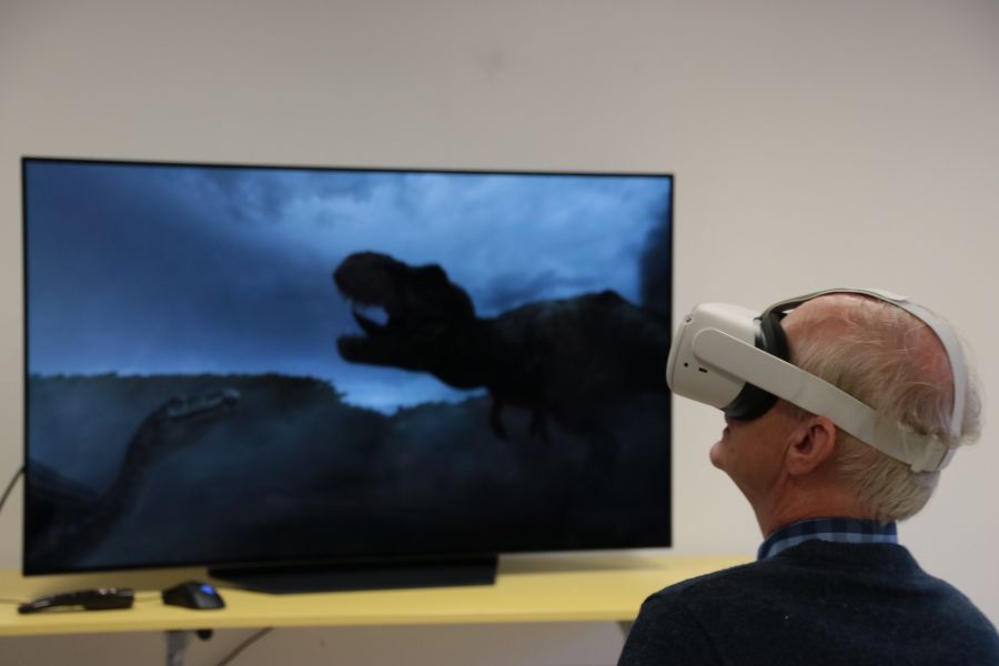 Na ekranie telewizora widać dinozaura, przed ekranem stoi mężczyzna w okularach do VR.