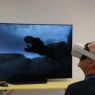 Na ekranie telewizora widać dinozaura, przed ekranem stoi mężczyzna w okularach do VR.