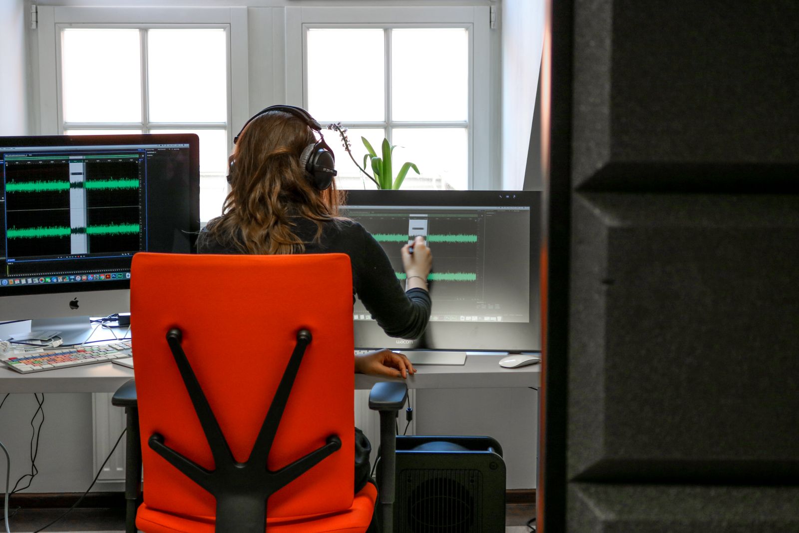 Młoda kobieta w słuchawkach siedzi tyłem do obiektywu na pomarańczowym krześle biurowym, przed nią dwa ekrany, na których wyświetla się ścieżka nagranego dźwięku.