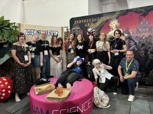 Studenci i studentki groznawstwa wraz z dr Joanną Pigulak i drem Marcinem Pigulakiem pozują do wspólnego zdjęcia na tle stoiska Fantastycznego UAM