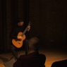 Wykonawca - Andrzej Grygier - siedzi na krześle w Sali Teatralnej, w przytłumionym świetle, grając na gitarze. Na pierwszym planie widać ciemne sylwetki publiczności z pierwszego rzędu.