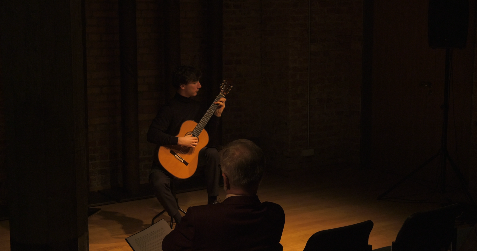 Wykonawca - Andrzej Grygier - siedzi na krześle w Sali Teatralnej, w przytłumionym świetle, grając na gitarze. Na pierwszym planie widać ciemne sylwetki publiczności z pierwszego rzędu.