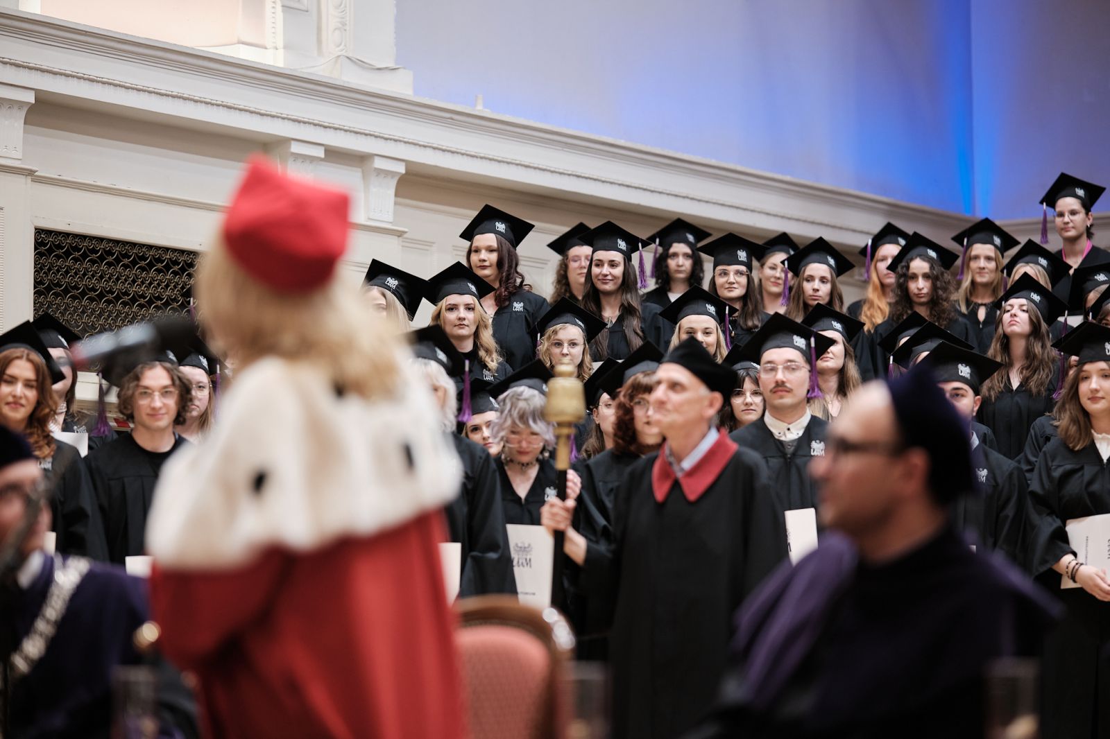 JM Rektor UAM zwraca się w przemowie do stojących za nią studentów w togach i biretach.