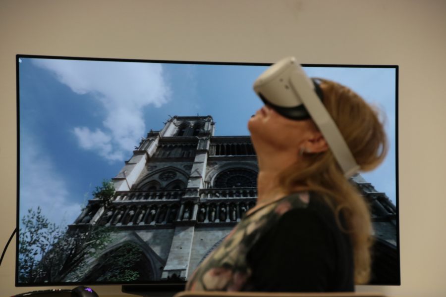Na ekranie telewizora widać katedrę Notre Dame w Paryżu, przed ekranem stoi kobieta w okularach VR, patrzy w górę.