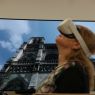 Na ekranie telewizora widać katedrę Notre Dame w Paryżu, przed ekranem stoi kobieta w okularach VR, patrzy w górę.