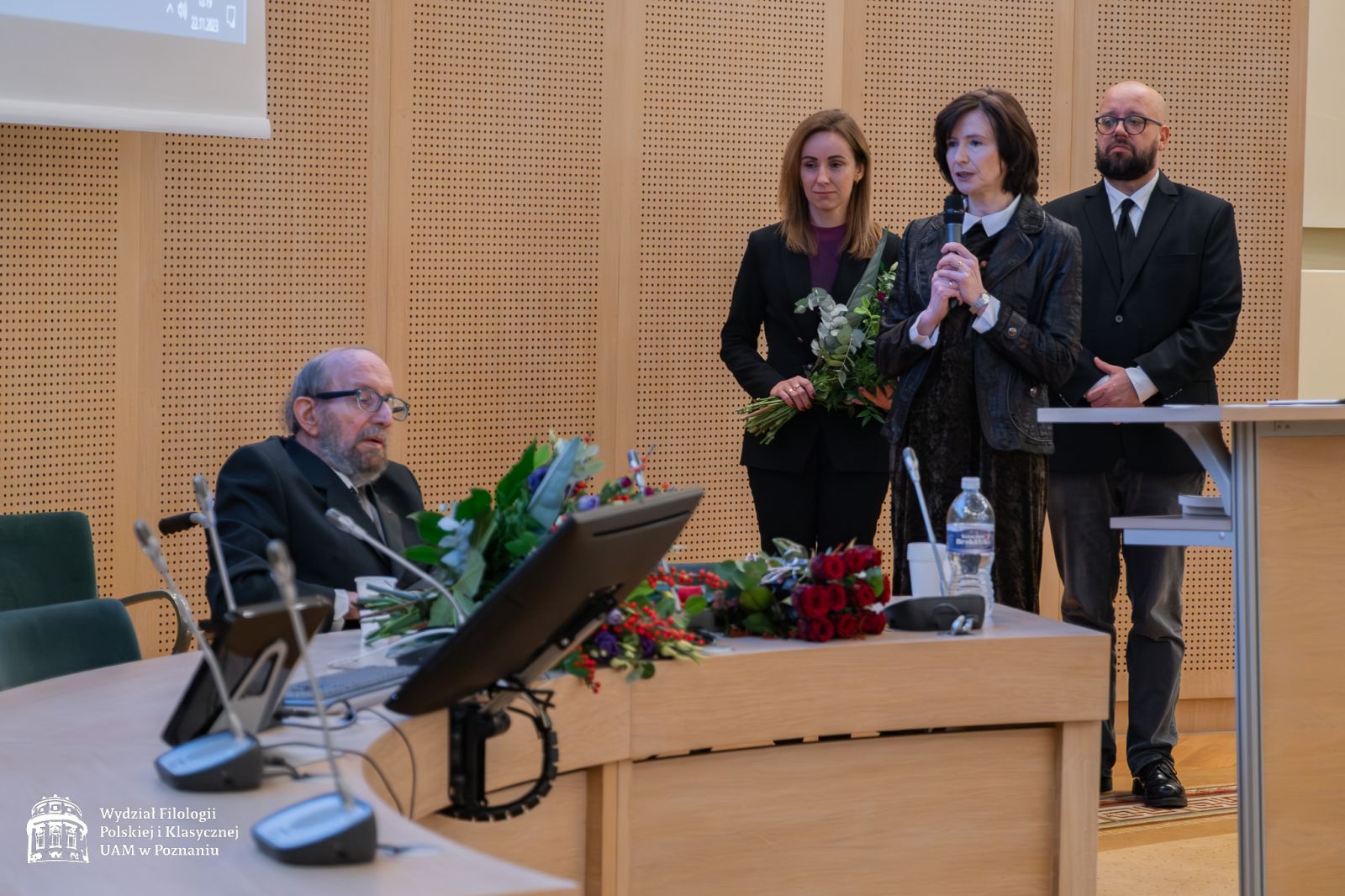 Dyrekcja IFP składa życzenia jubilatowi - przy mikrofonie stoi prof. Elżbieta Winiecka, za nią dr Sylwia Karolak z bukietem kwiatów w rękach, jeszcze dalej prof. Marek Osiewicz.