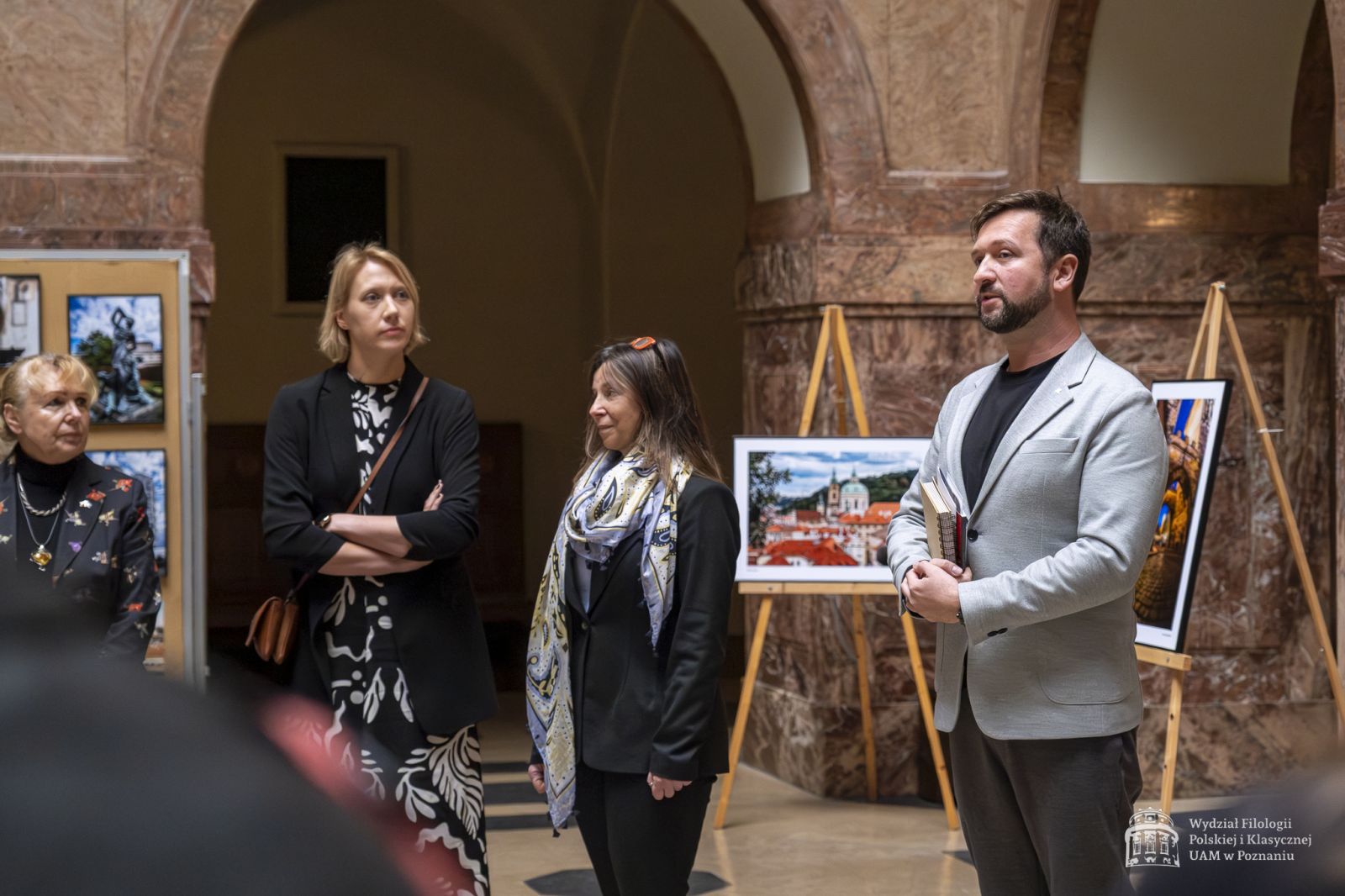 Dyrektor IFS dr hab. Patryk Borowiak przemawia w holu głównym, obok niego stoją: dr Joanna Brodniewicz, dr Urszula Kowalska-Nadolna i konsul honorowa Czech Renata Mataczyńska. Za nimi widać sztalugi z wystawą.