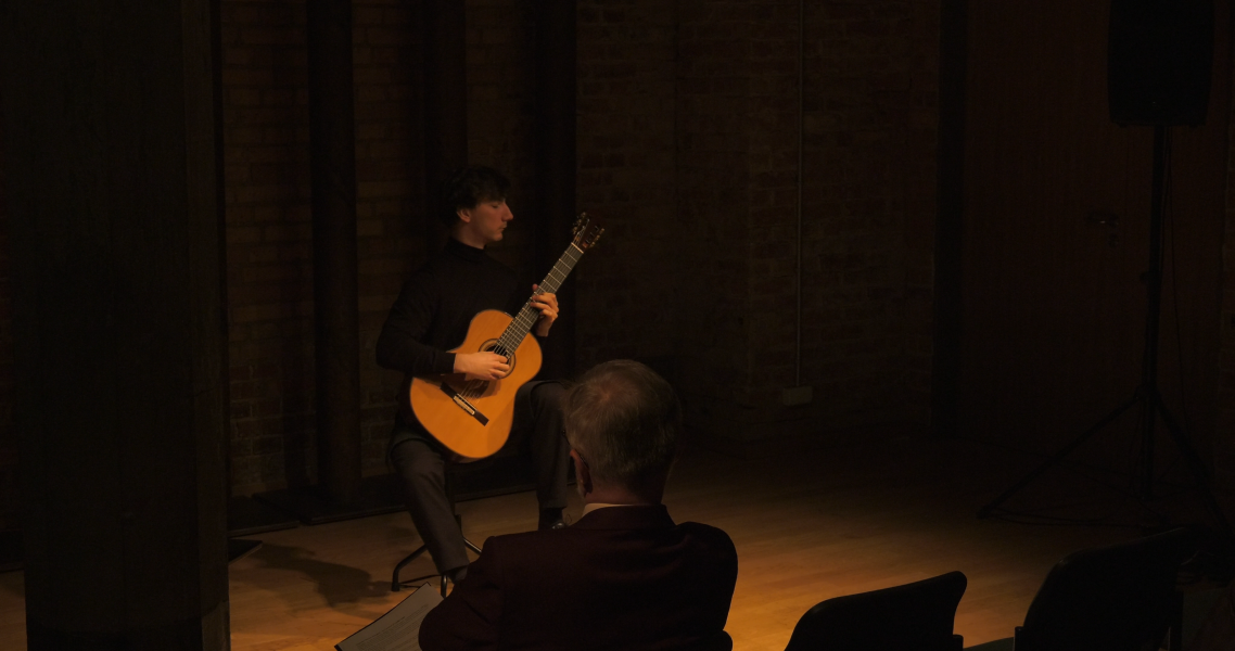 Wykonawca - Andrzej Grygier - siedzi na krześle w Sali Teatralnej, w przytłumionym świetle, grając na gitarze. Na pierwszym planie widać ciemne sylwetki publiczności z pierwszego rzędu.