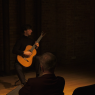 Wykonawca - Andrzej Grygier - siedzi na krześle w Sali Teatralnej, w przytłumionym świetle, grając na gitarze. Na pierwszym planie widać ciemne sylwetki publiczności z pierwszego rzędu.