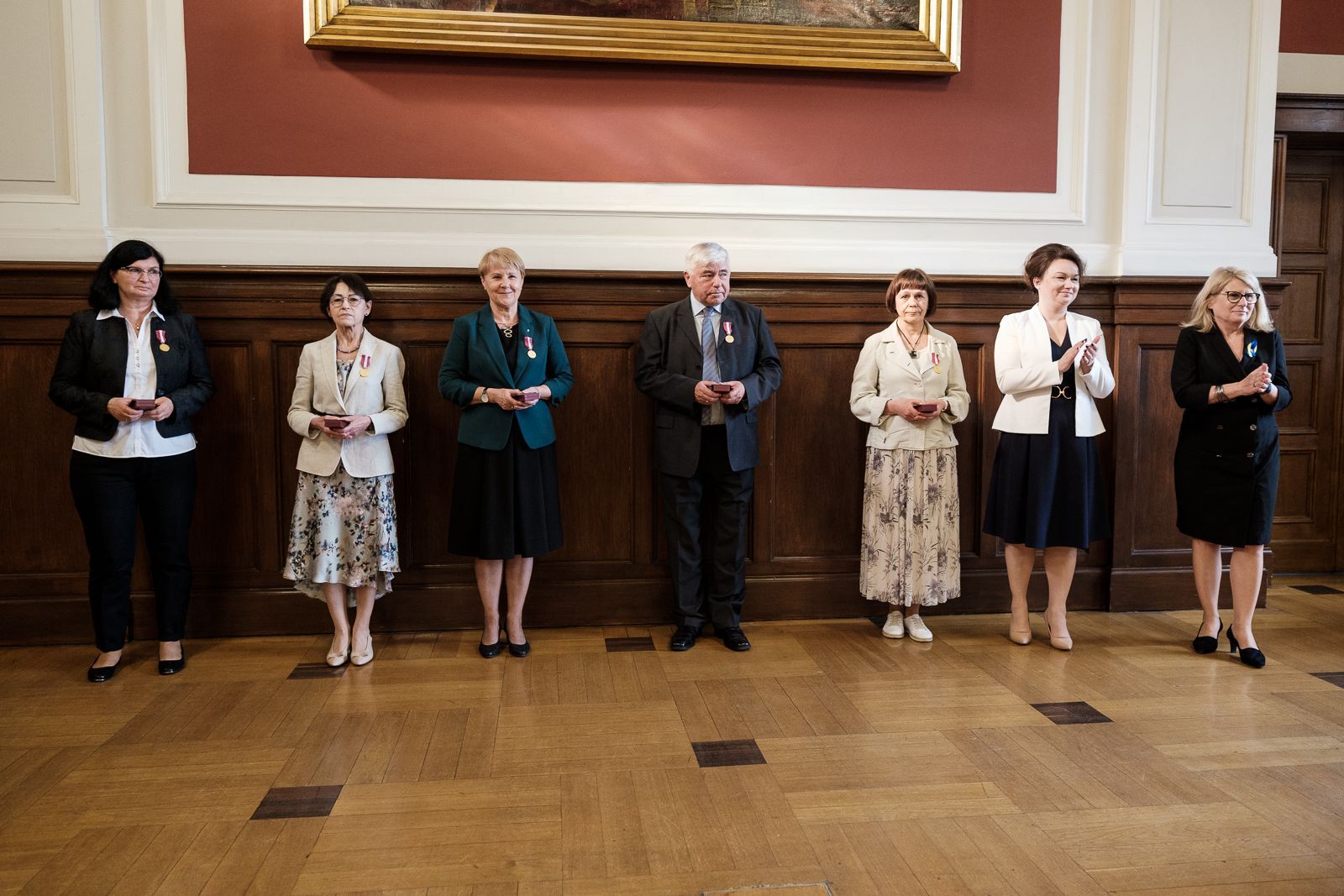 Na tle ściany wyłożonej drewnem stoi siedem osób, pięć z nich na piersi ma medal, dwie klaszczą.