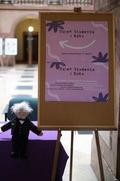 W holu głównym Collegium Maius stoi sztaluga z plakatami reklamującymi Dzień Studenta I roku, obok niej na stole pokrytym fioletowym suknem siedzi pacynka Aleksandra Fredry.