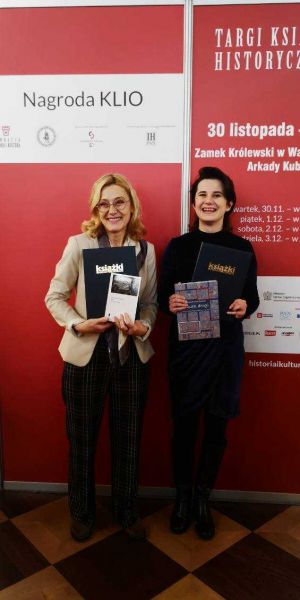 Dwie kobiety stoją na tle ścianki reklamującej Targi Książki Historycznej w Warszawie. Obie trzymają w rękach książki i śmieją się do obiektywu.
