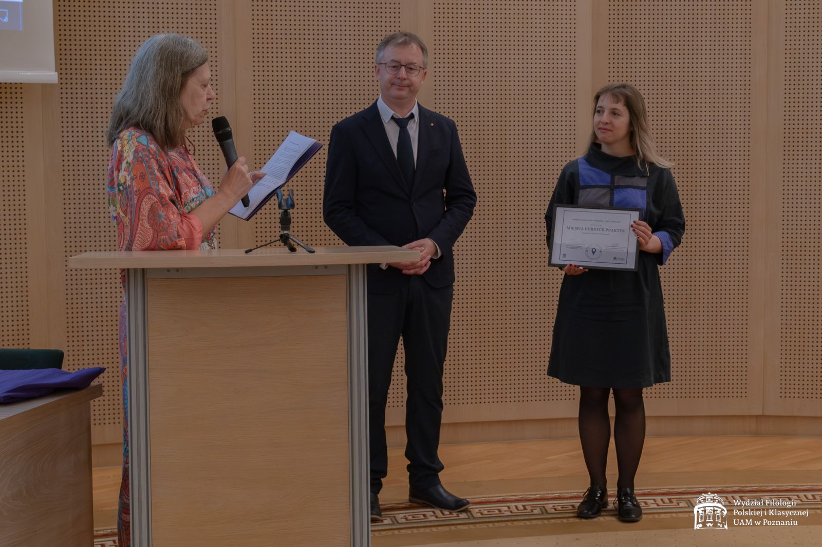 Prof. Anna Gawarecka odczytuje laudację, obok niej stoi Dziekan WFPiK, a dalej młoda kobieta z dyplomem 