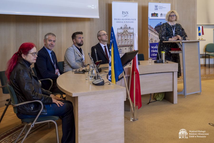 Prof. Ewa Szperlik przemawia przy mównicy, obok niej za stołem prezydialnym siedzą: dyrektorka Teatru im. Fredry w Gnieźnie Joanna Nowak, prof. Sanjin Kodrić, dr hab. Patryk Borowiak, prof. UAM dr hab. Krzysztof Skibski. Przed stołem prezydialnym ustawiono flagę Polski i Bośni i Hercegowiny.