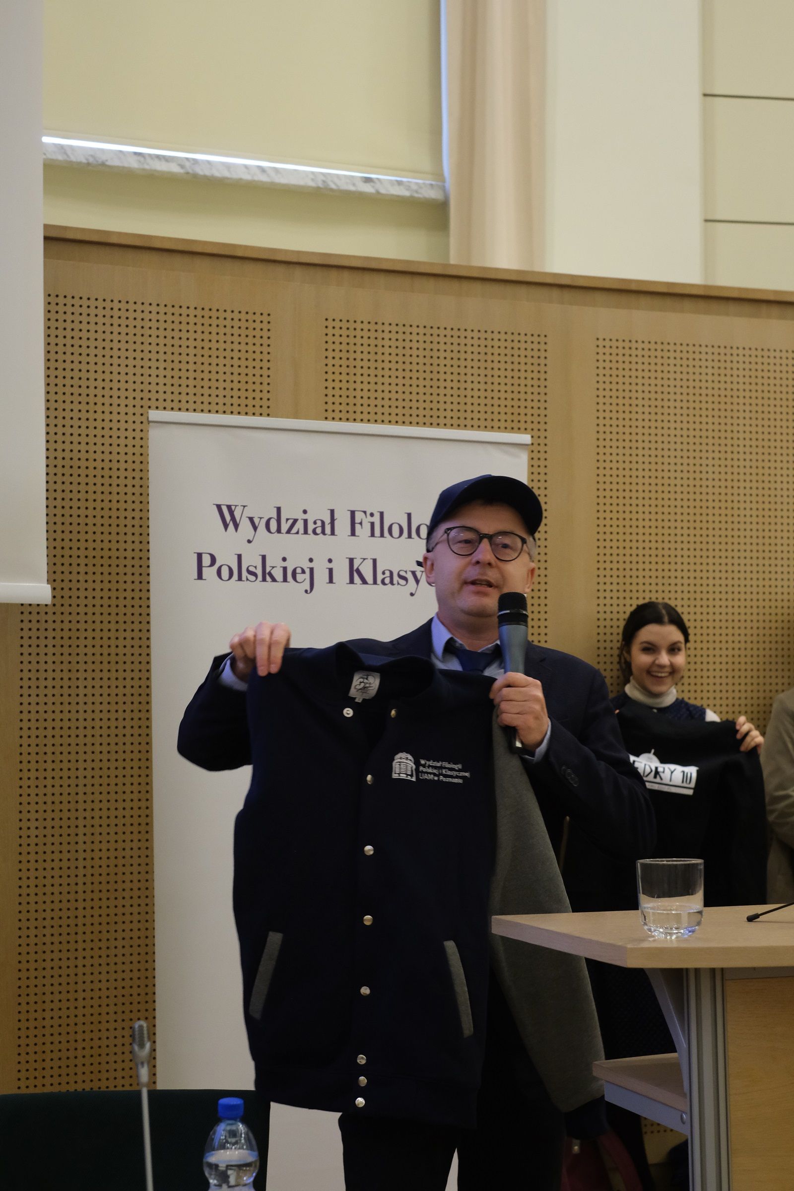 Dziekan WFPiK stoi przy mównicy i przemawia do mikrofonu, prezentuje bluzę wydziałową, na głowie ma czapkę z daszkiem, również z oznakowaniem wydziałowym.