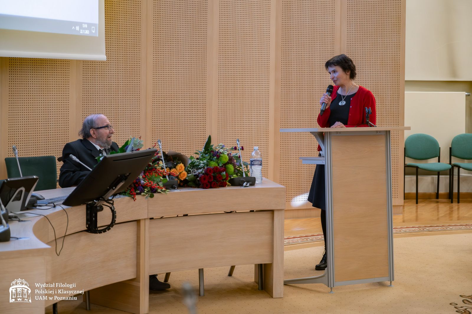 Przy mównicy stoi kobieta w średnim wieku, ubrana w ciemną sukienkę i czerwony sweterek, krewna prof. Lewandowskiego, przemawia do mikrofonu.