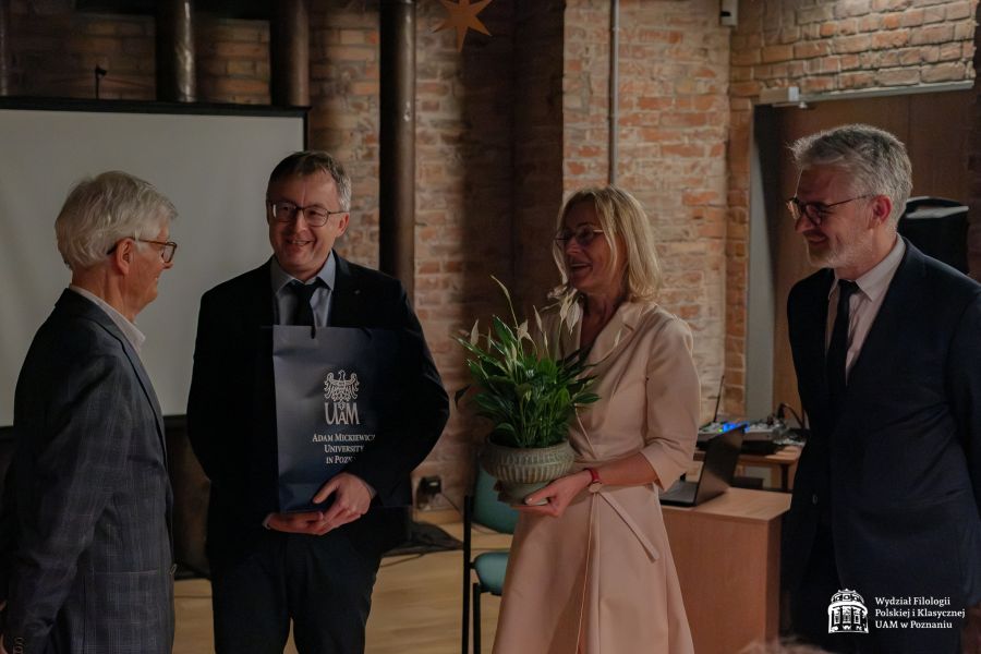 Grono dziekańskie gratuluje prof. Przychodniakowi, dziekan Mizerkiewicz trzyma prezent w torbie UAM, a prof. Krystyna Pieniążek-Marković trzyma doniczkę z kwiatami.