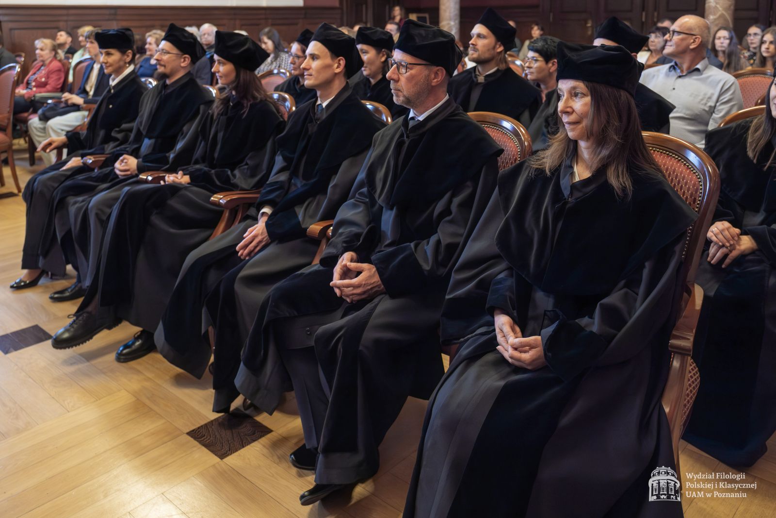 Doktoranci w czarnych togach siedzą w dwóch rzędach na krzesłach.